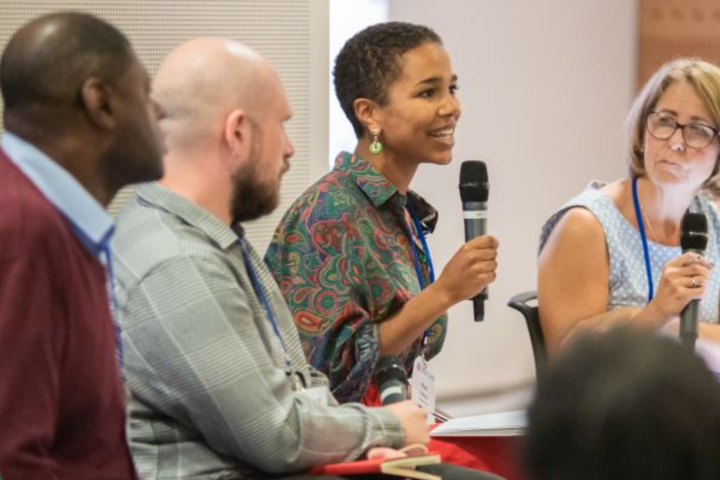 Panel of four speakers sat down, with the two to the right holding microphones, and the person second from right speaking to a crowd