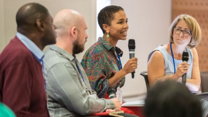 Panel of four speakers sat down, with the two to the right holding microphones, and the person second from right speaking to a crowd