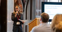 Female speaker with a microphone and notes addresses a room, with a screen behind them with logos from Big Society Capital and Snowball