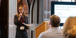 Female speaker with a microphone and notes addresses a room, with a screen behind them with logos from Big Society Capital and Snowball