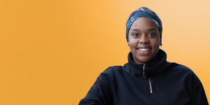 Woman wearing a head scarf and dark clothing smiling against a yellow background