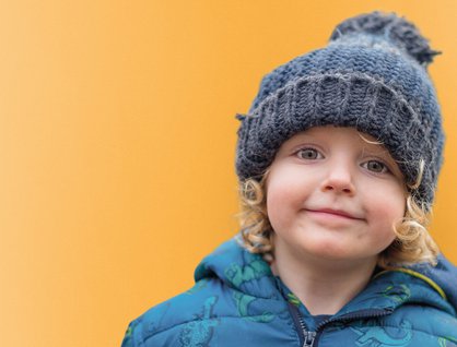Blonde child wearing a blue puffer coat and beanie in front of a yellow background