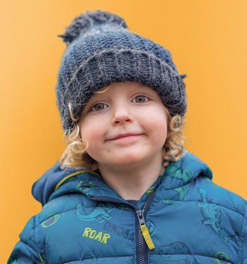 Blonde child wearing a blue puffer coat and beanie in front of a yellow background