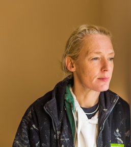 Woman wearing a black fleece with paint marks looking out of a window