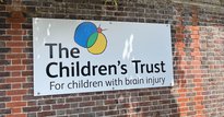 White sign for the Children's Trust affixed to a brick wall, with trees behind