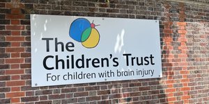 White sign for the Children's Trust affixed to a brick wall, with trees behind