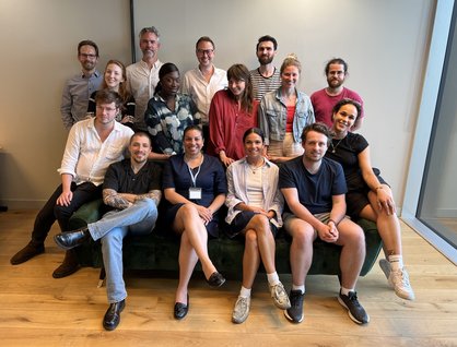 Group of 15 people posing around a sofa in an office space, smiling at the camera