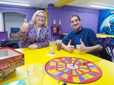 Two people sat around a table in a purple room, playing a board game and smiling at the camera