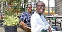 A man with glasses and a woman in a plaid shirt sitting on a bench in a garden smiling