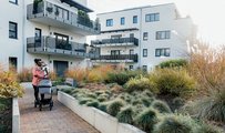 Parent walking with baby and stroller through landscaped courtyard of a modern residential apartment complex.