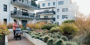 Parent walking with baby and stroller through landscaped courtyard of a modern residential apartment complex.