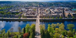 Birds eye view of a river, with lots of trees on one side and buildings on the other, with a road bridge going across