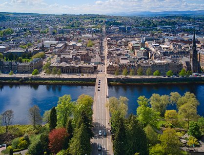 Birds eye view of a river, with lots of trees on one side and buildings on the other, with a road bridge going across
