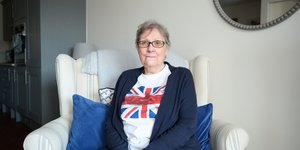 Resident at Uxbridge United Welfare Trust pictured in her apartment. She is sitting on a chair, wearing a tshirt with a union flag on it.